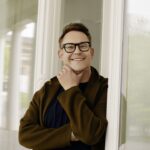 Lächelnde Person mit Brille steht an Fenster.